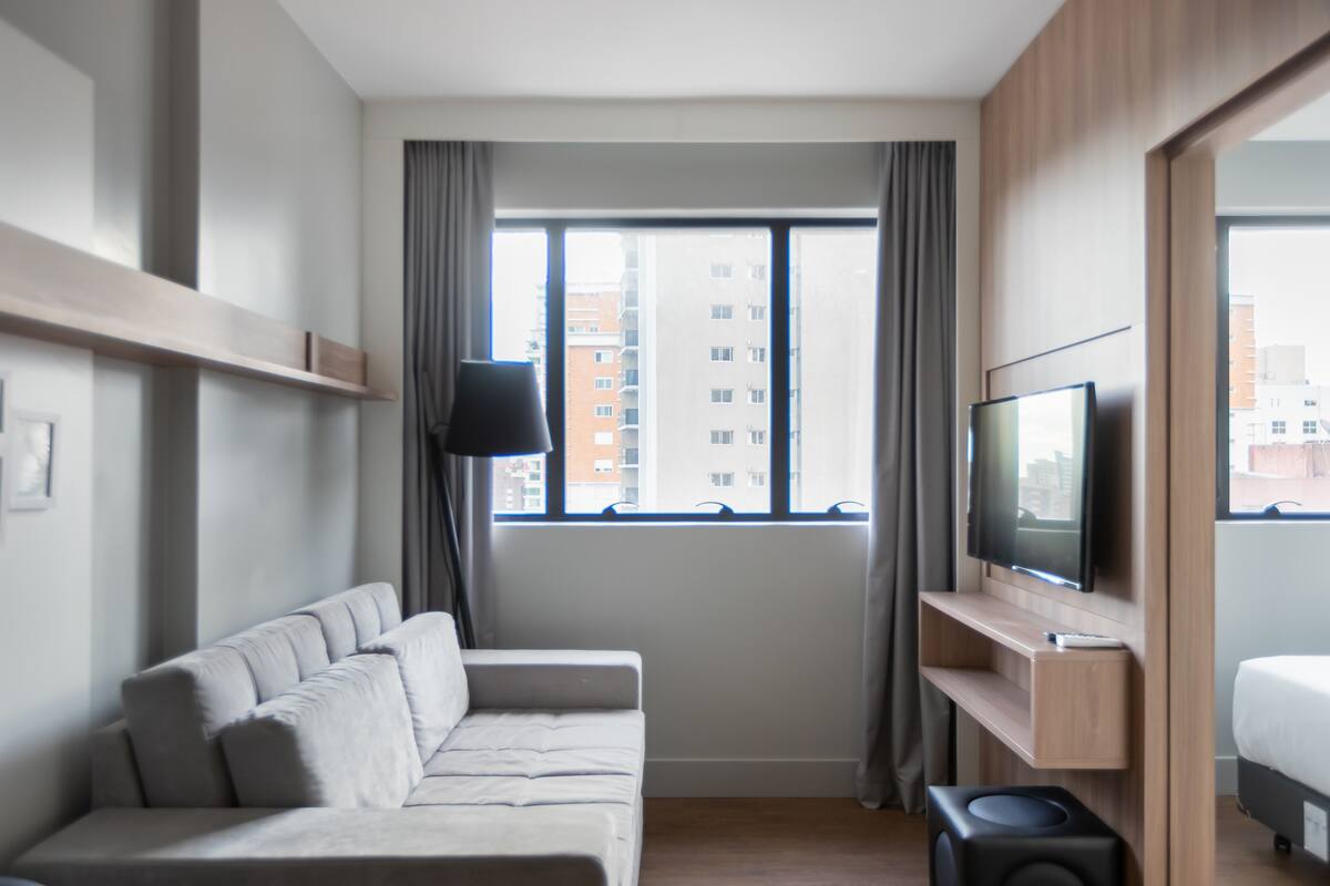 Une chambre d’hôtel moderne équipée d’un canapé, d’une télévision, d’une petite table et d’une fenêtre donnant sur des immeubles.