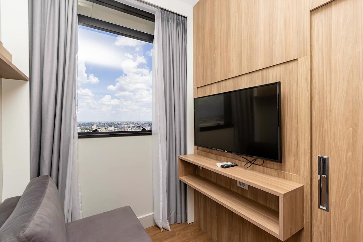 Un salon moderne avec un téléviseur à écran plat fixé à un mur en bois, des rideaux gris et une vue sur la ville par une grande fenêtre.