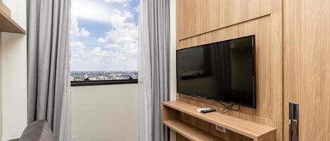 Una sala de estar moderna con un televisor de pantalla plana montado en una pared de madera, cortinas grises y vistas de la ciudad a través de un ventanal grande.