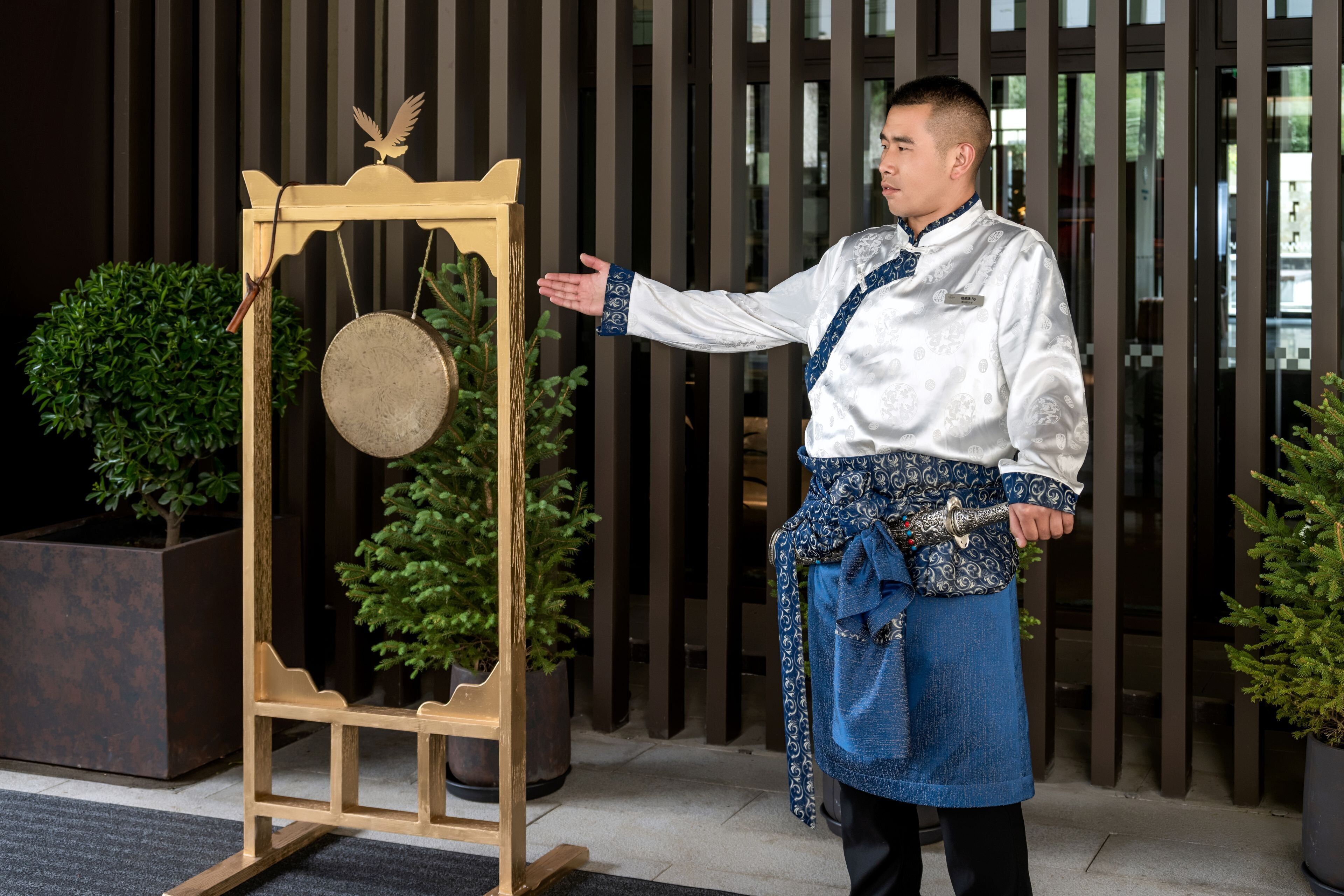 A person standing next to a wooden display with a circular object and a bird figure, in front of a building with vertical siding and potted plants.