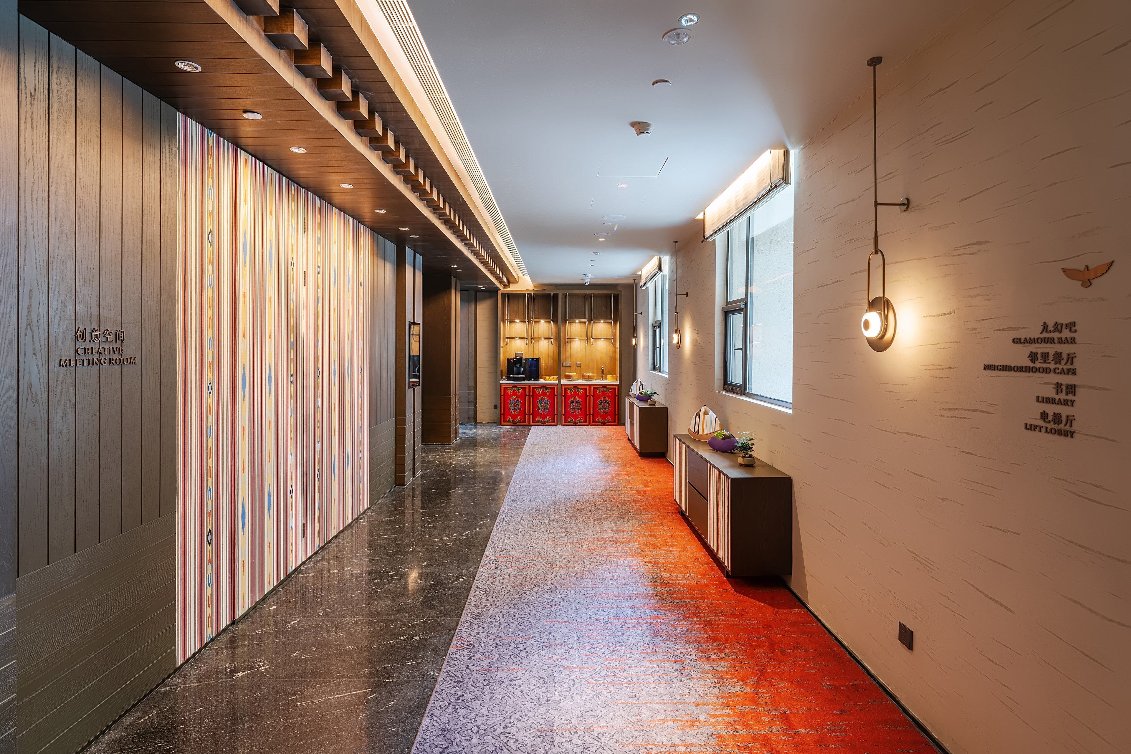 Un couloir d’hôtel moderne avec un mur en bois sur la gauche, une moquette rouge et un luminaire fixé au mur sur la droite.