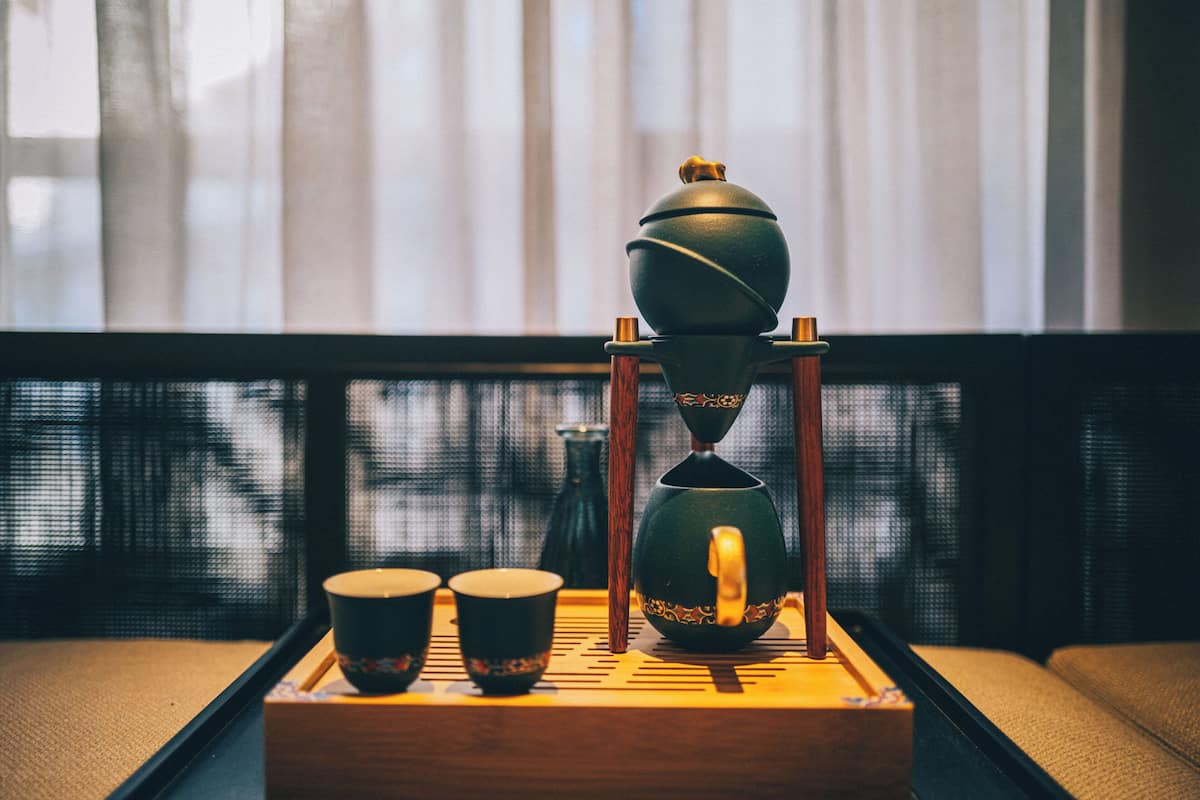 Un service à thé traditionnel, posé sur un plateau avec deux tasses et une théière sur un présentoir, est placé sur une table en bois près d’une fenêtre ornée de rideaux légers.