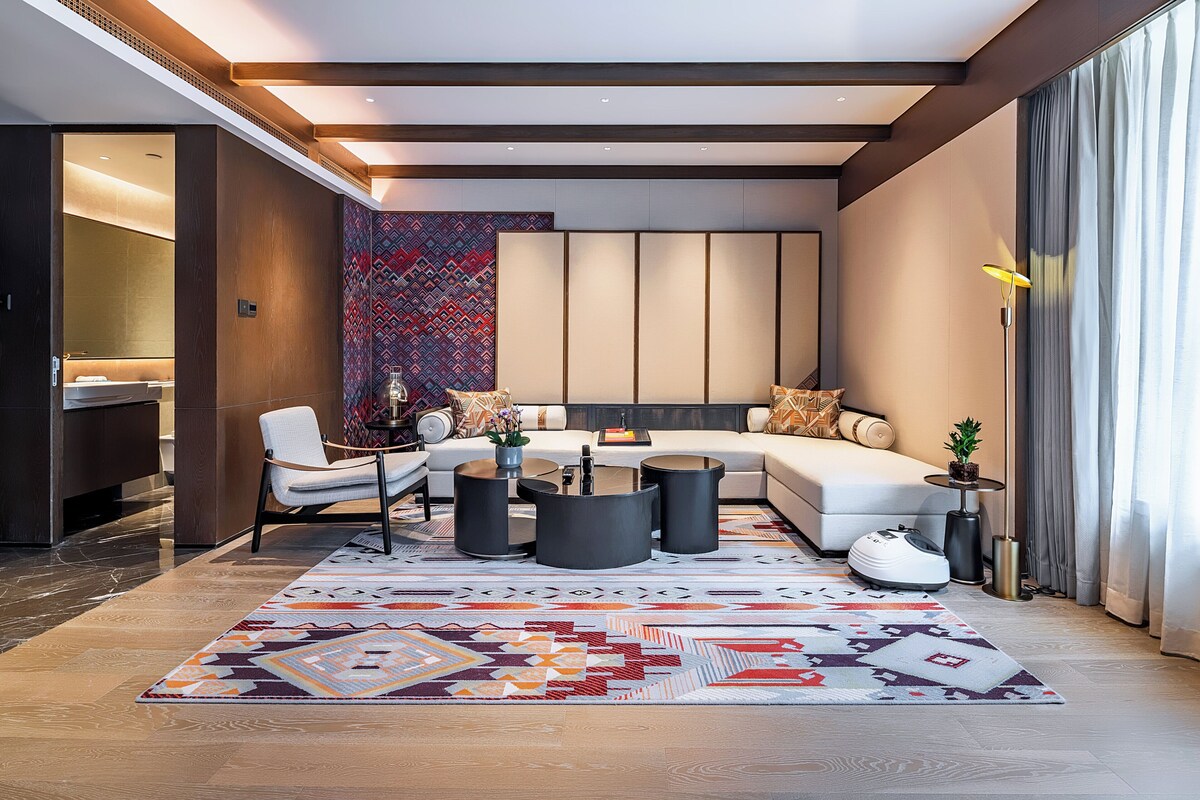 Ein modernes Wohnzimmer mit einer Eckcouch, zwei runden Couchtischen, einem gemusterten Teppich und einem kleinen Beistelltisch mit einer Pflanze.