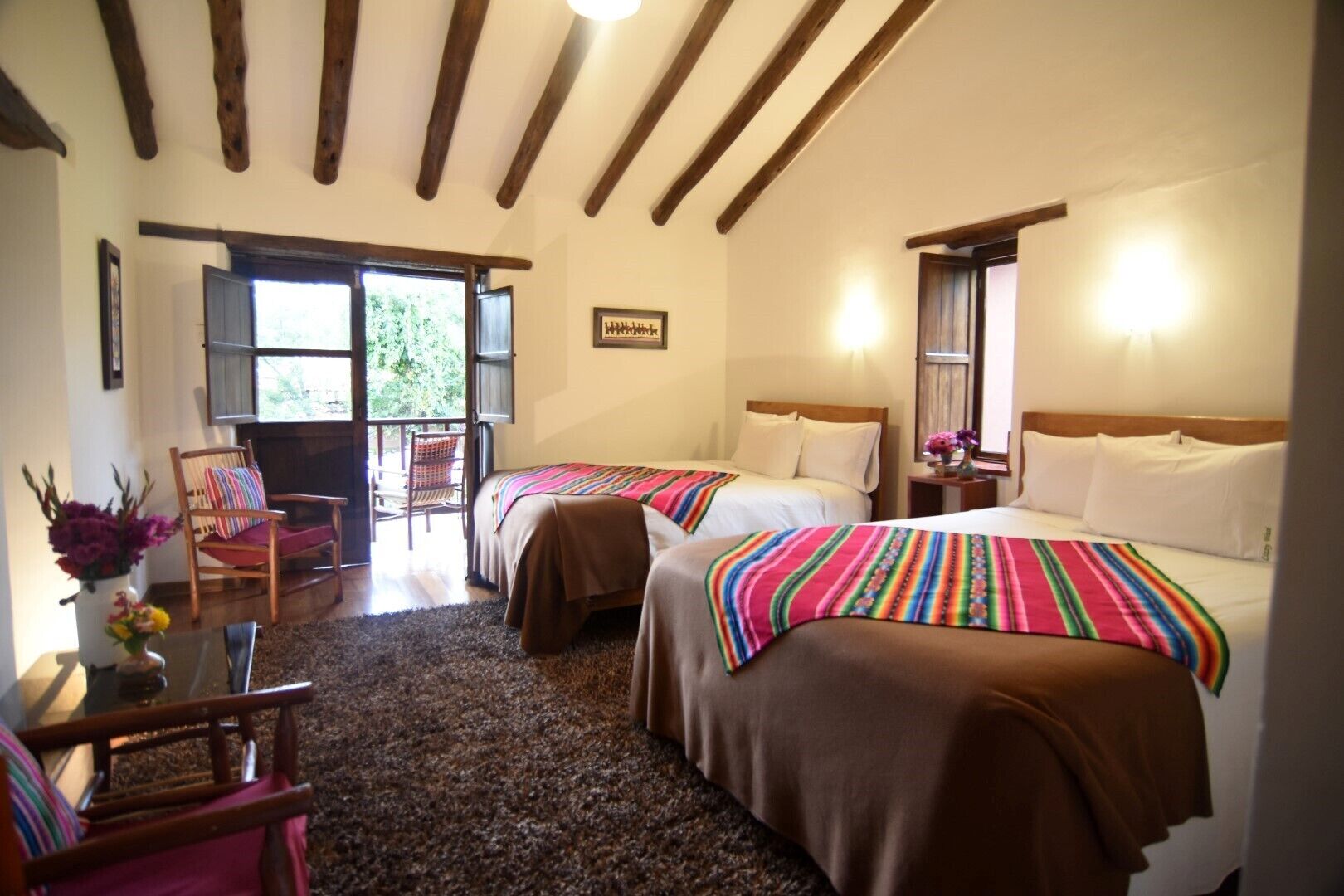 A hotel room with two beds, a wooden ceiling, a chair, a table, and a view through a door.
