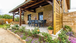 Un edificio de piedra con un patio cubierto, mobiliario exterior y macizos de flores.