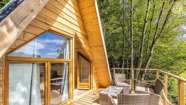 Une cabane en bois dotée de grandes fenêtres, d’une terrasse avec des meubles en osier et offrant une vue sur une forêt.