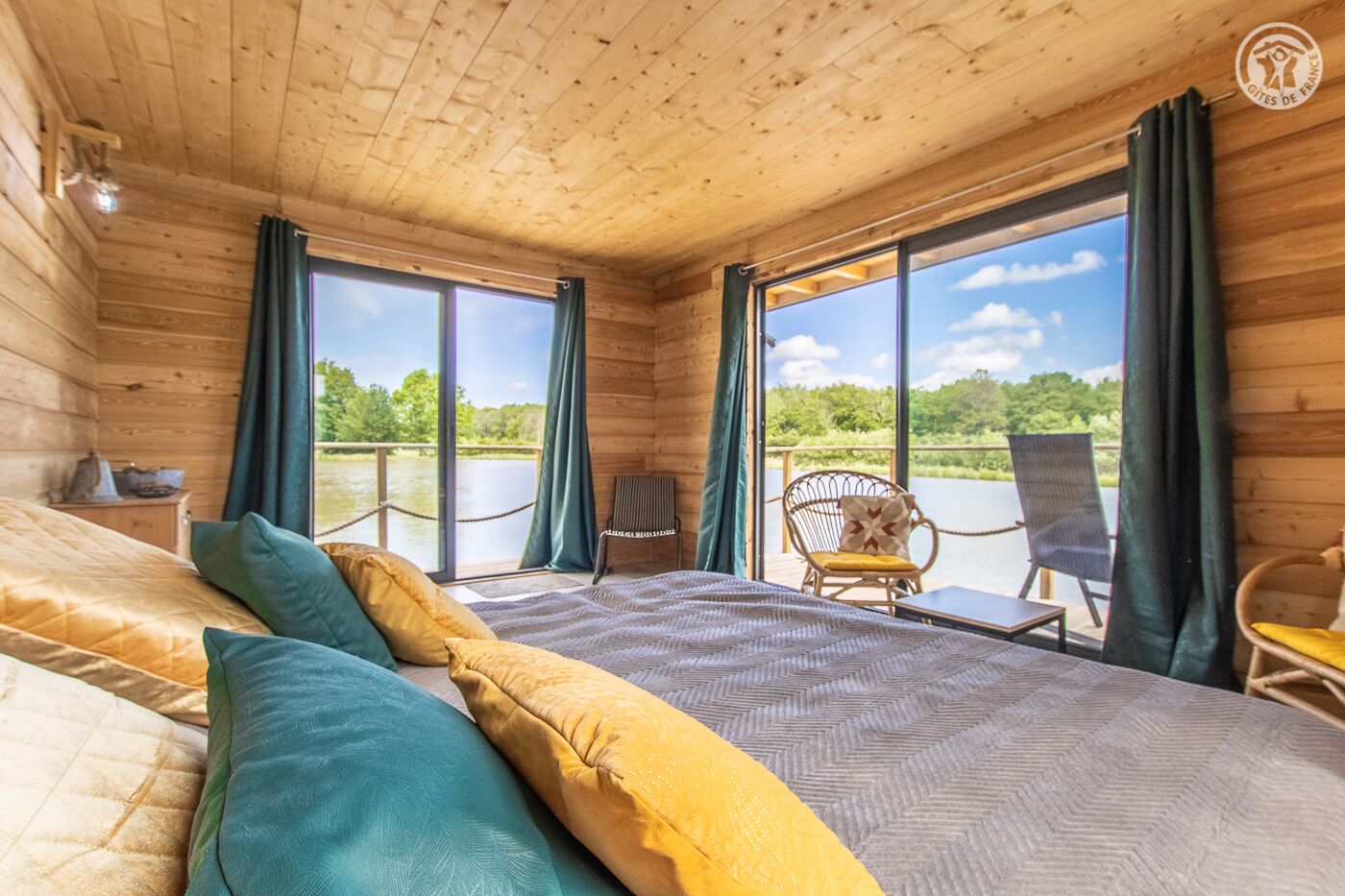 Une pièce de cabane en bois comprenant un lit, deux chaises et de grandes fenêtres donnant sur une étendue d’eau.