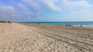 A wide sandy beach with visible tire tracks, a clear sky, and the ocean in the background. - Coco Sands Studios (Cocoa Beach)
