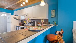 A kitchen with white cabinets, a blue accent wall, a wooden ceiling, and a bar area with stools. - Coco Sands Studios (Cocoa Beach)