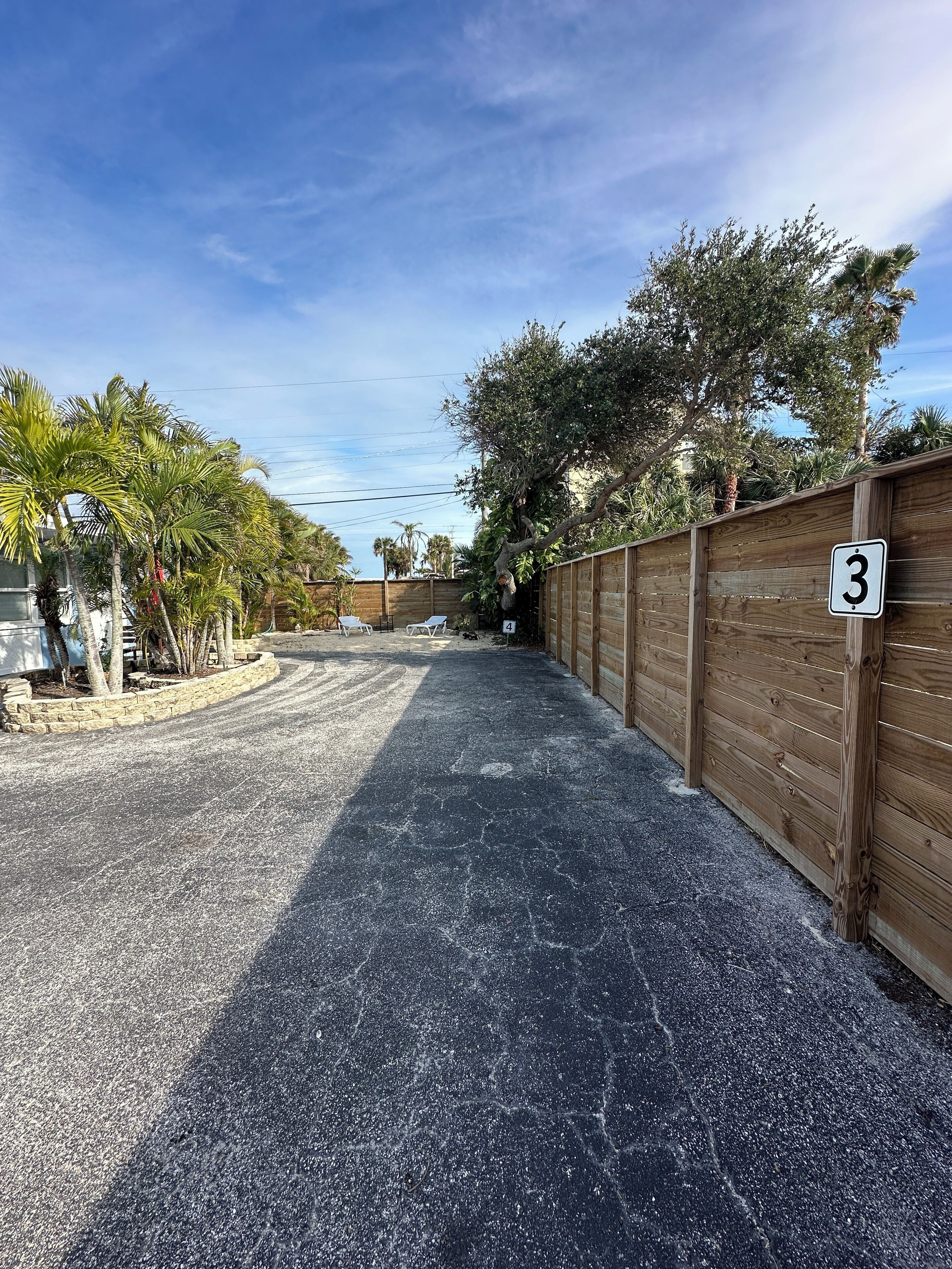 A driveway leading to a wooden fence with a number '3' sign, surrounded by palm trees and a clear sky.