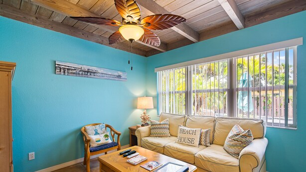 A cozy living room with a blue wall, a wooden ceiling fan, a beige sofa with a 'Beach House' pillow, a wooden coffee table, and a window with white blinds. - Coco Sands Studios (Cocoa Beach)