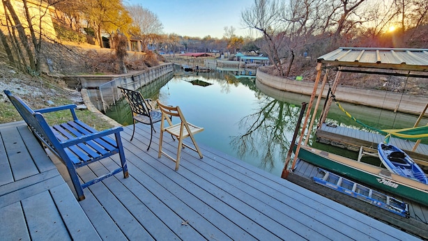A wooden deck with a bench and chairs overlooking a calm body of water, a dock with boats, and a building with a green roof. - Huge 4 bedroom + loft lakehouse with private dock - 27 min north of Sundance Sq  (Fort Worth)