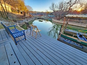 Una terraza de madera con banco y sillas que da a una masa de agua tranquila, un muelle con barcos y un edificio con techo verde.
