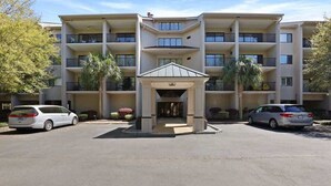 A multi-story residential building with a central entrance, balconies on each floor, and a parking lot with cars. - Port Royal Village 302 - Walk to Beach (Hilton Head Island)