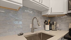 A kitchen with a stainless steel sink, white cabinets, and a grey tiled backsplash. - Port Royal Village 302 - Walk to Beach (Hilton Head Island)