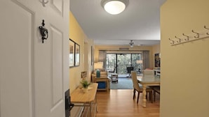 A well-lit hallway leading to a dining area with a table and chairs, a ceiling fan, and a view of the outdoors through large windows. - Port Royal Village 302 - Walk to Beach (Hilton Head Island)