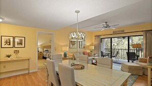 A spacious living room with a dining table, sofas, and a ceiling fan. There is a balcony with outdoor seating and a view of trees. - Port Royal Village 302 - Walk to Beach (Hilton Head Island)