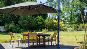 An outdoor patio with a table and chairs under a large umbrella, surrounded by greenery.