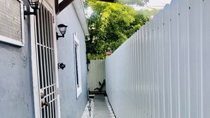 A narrow concrete pathway flanked by a white fence and a building with a metal gate and a door.