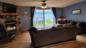 A living room with a stone fireplace, a large sofa, a television, and a view of a lake through a glass door. - Lakefront cottage on Secluded Upper Herring Lake (Frankfort)