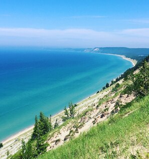 A coastal landscape with a clear view of the ocean, a sandy beach, and a grassy hillside. - Lakefront cottage on Secluded Upper Herring Lake (Frankfort)
