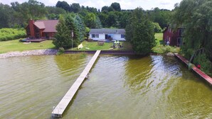A lakeside property with a dock extending into the water, surrounded by lush greenery and a large house with a deck. - Lakefront cottage on Secluded Upper Herring Lake (Frankfort)