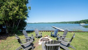 A lakeside deck with Adirondack chairs, a fire pit, and a dock extending into the water. - Lakefront cottage on Secluded Upper Herring Lake (Frankfort)