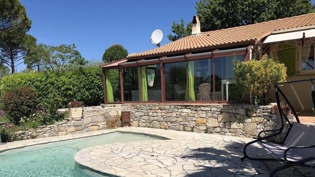 A swimming pool with stone tiles, a stone wall, and a covered patio area with a chair. - Gîte de Font du Roux (FLAYOSC)