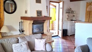 A living room with a sofa, a wooden coffee table, a fireplace, and a kitchen area visible through an open door. - Gîte de Font du Roux (FLAYOSC)