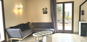 A living room with a grey sofa, a small table, a teddy bear, a wall-mounted light fixture, and a sliding glass door leading to an outdoor area.