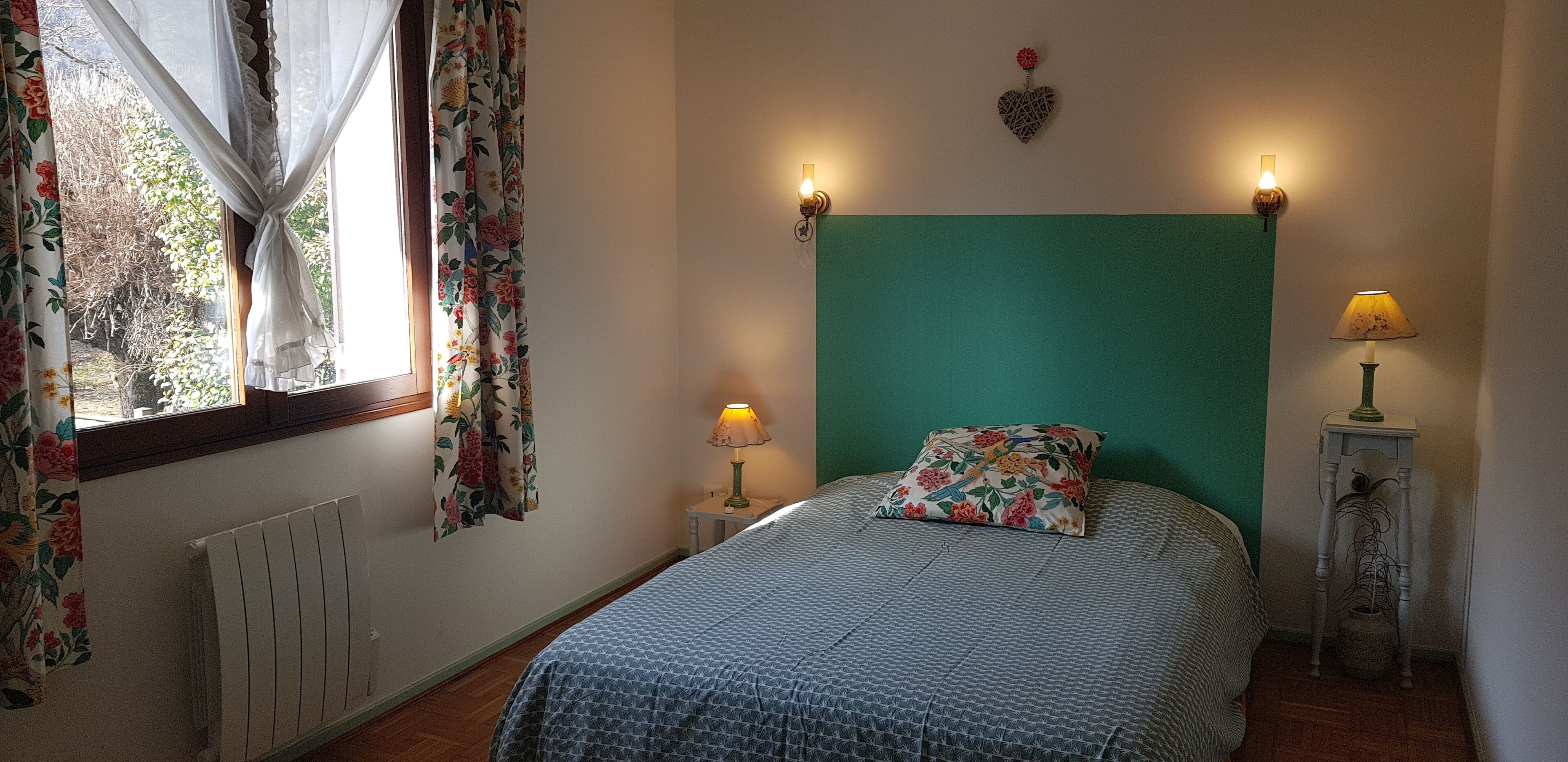 Ein Schlafzimmer mit einem Bett, floralen Vorhängen, einem grünen Kopfende, zwei Nachttischlampen und einer Heizung.