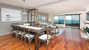 A spacious living area with a wooden dining table and chairs, a glass shelving unit, and a large window offering a view of the sea. - High End Oceanfront Home, a 2 min walk to Beach, Shops, Dining & More in Solana (Solana Beach)