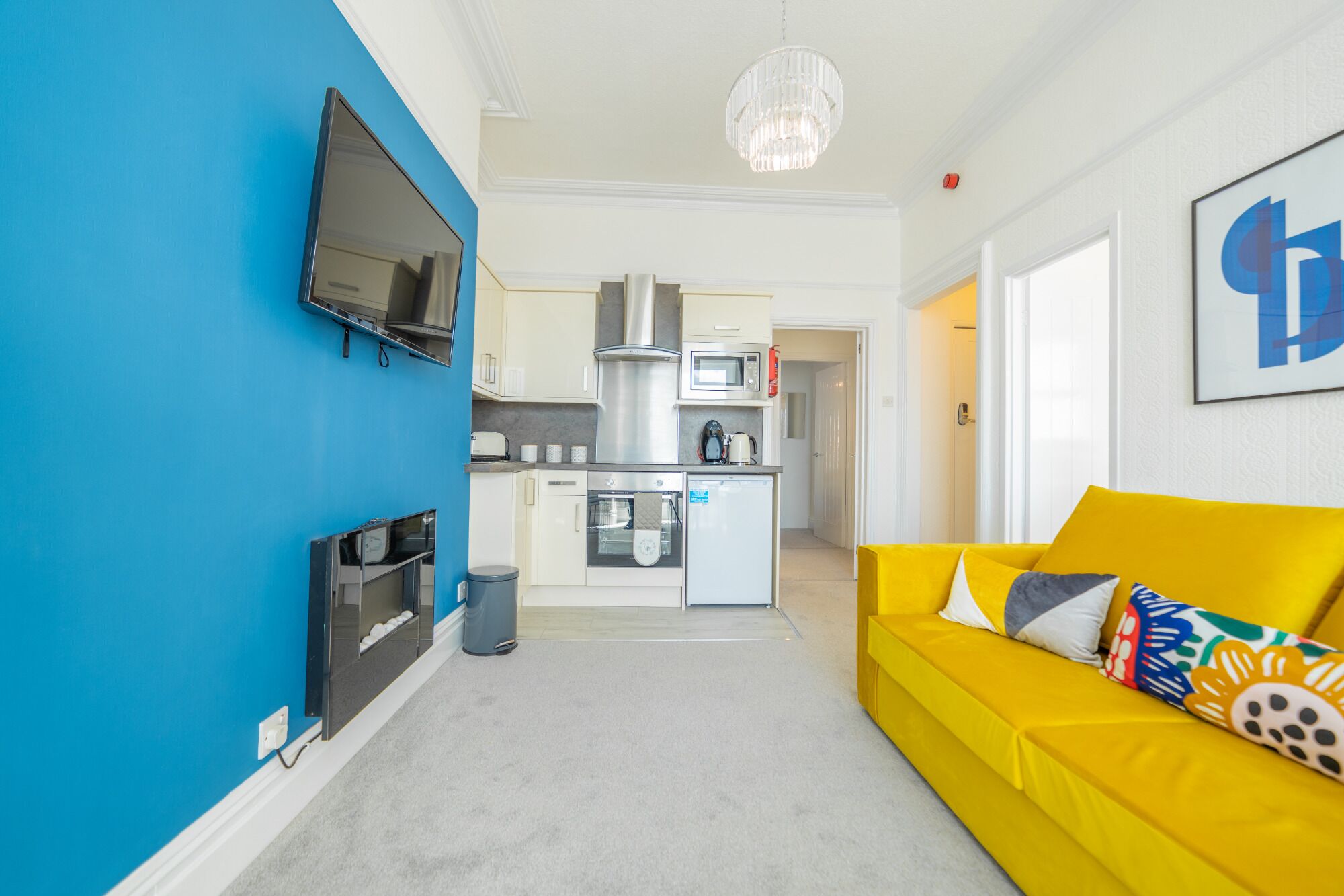 Un espacio habitable compacto con kitchenette, un sofá con cojines de colores, un televisor de pantalla plana montado en una pared azul y una araña.