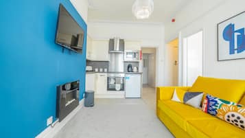 Un espacio habitable compacto con kitchenette, un sofá con cojines de colores, un televisor de pantalla plana montado en una pared azul y una araña.