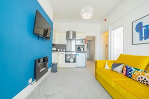 Ein kompakter Wohnbereich mit Küchenzeile, einer Couch mit bunten Kissen, einem an einer blauen Wand angebrachten Flachbildfernseher und einer Deckenleuchte.