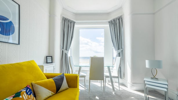 A bright room with a yellow sofa, a glass table, and a view of the beach. - Highcliffe Seaview Suite (Blackpool)
