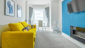 A modern living room with a yellow sofa, a flat-screen TV, and a view to the outside. - Highcliffe Seaview Suite (Blackpool)