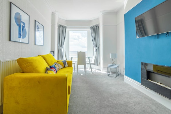 A modern living room with a yellow sofa, a flat-screen TV, and a view to the outside.