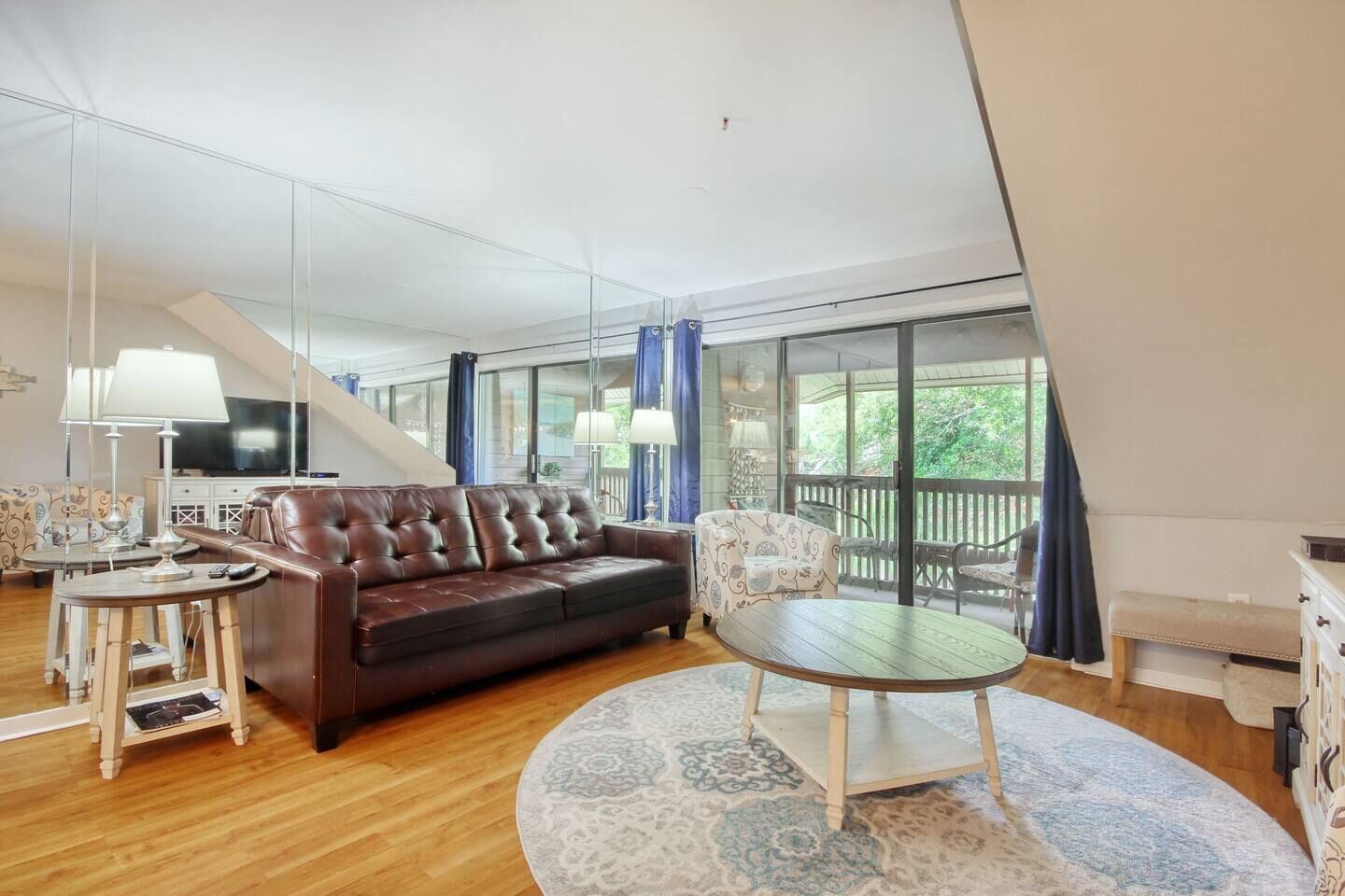 A modern living room with a leather sofa, a round table, and a TV. There is a balcony with a view of greenery.