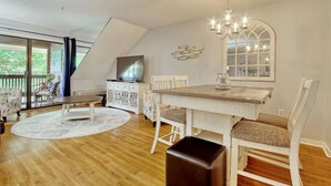 A spacious loft bedroom with a large window, a chandelier, a dining table, and a TV on a white cabinet. - LakeViews-Quaint/Quiet-Wlk 2 Beach-Kingston Plantation-Gated Community (Myrtle Beach)