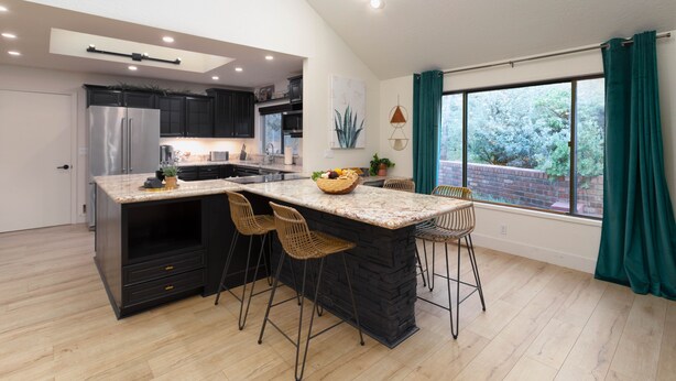 A modern kitchen with a central island, dark cabinetry, and a large window with curtains.