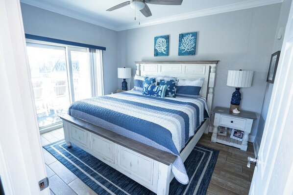 A bedroom with a large bed, two bedside tables, a ceiling fan, and a view of the outside.