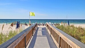 Une promenade en bois menant à une plage où se trouvent des parasols et un drapeau.