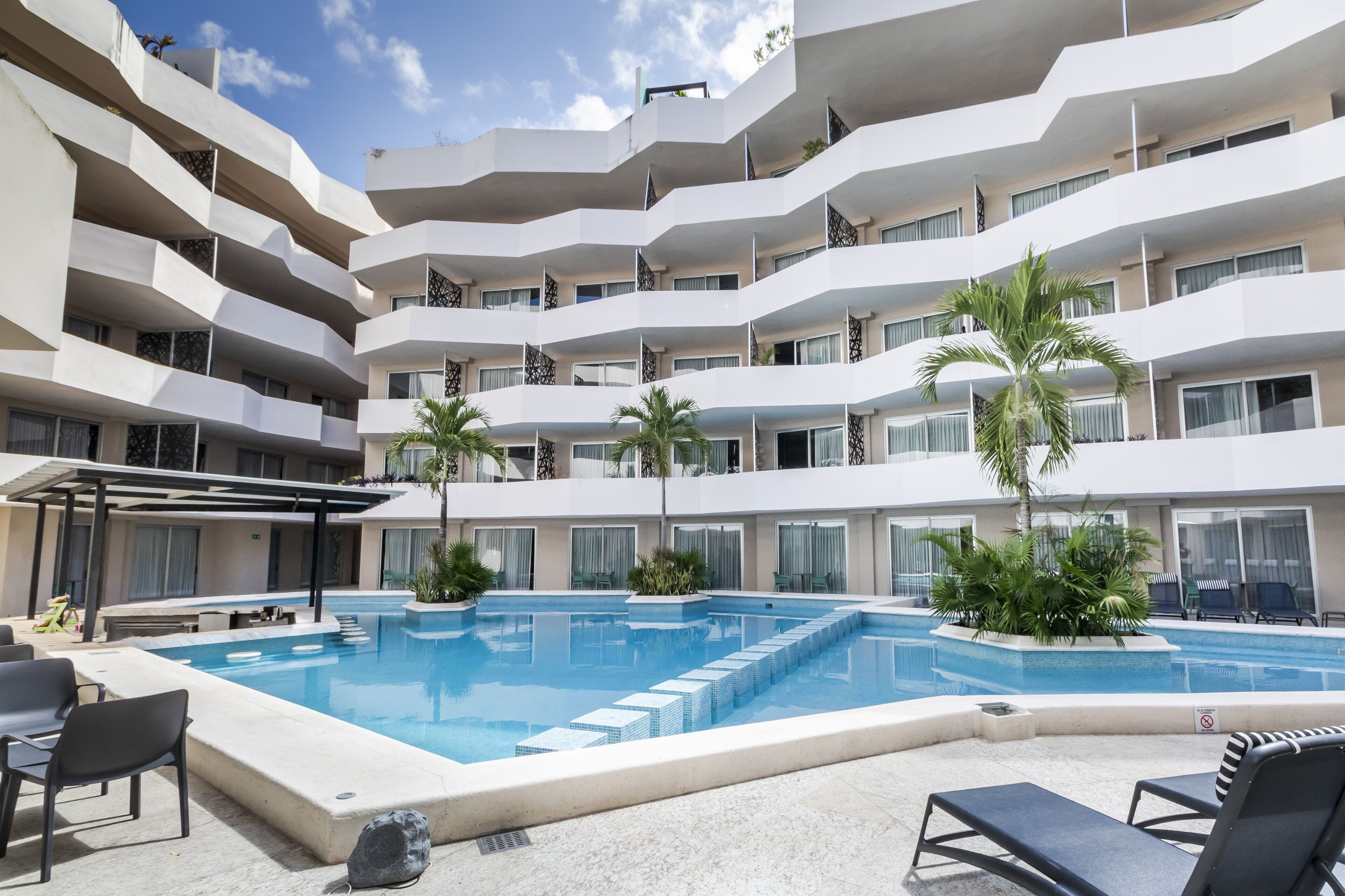A multi-story hotel with a central swimming pool surrounded by lounge chairs and palm trees.
