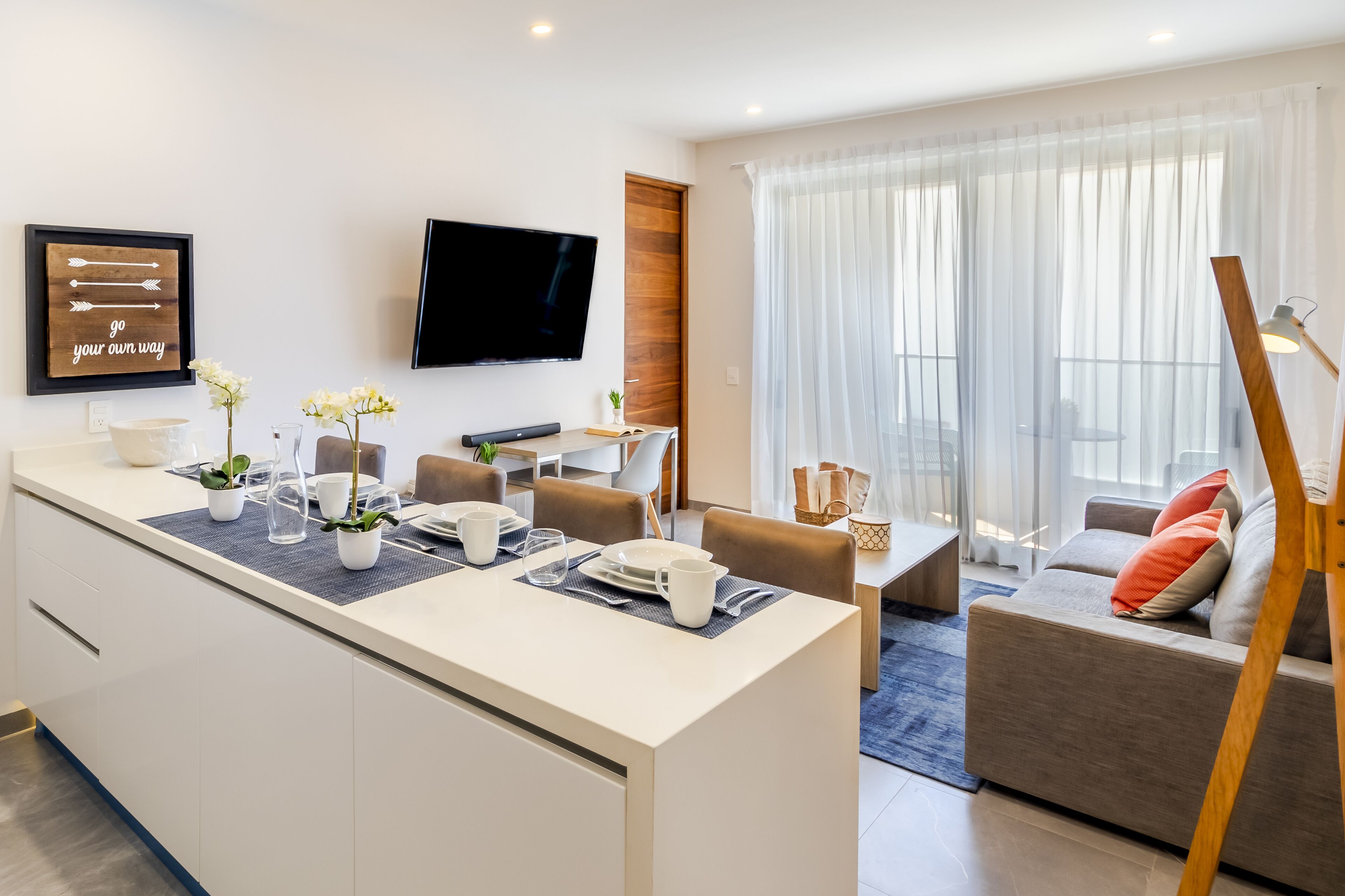 Una habitación de hotel moderna con zona de comedor, sofá y televisor en la pared.