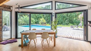 A spacious room with a large table and chairs, a view of a swimming pool, and a wooden deck outside.