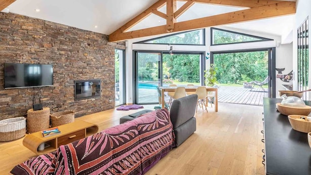 A modern living room with a stone wall, wooden beams, a large TV, and a view of a pool and garden.