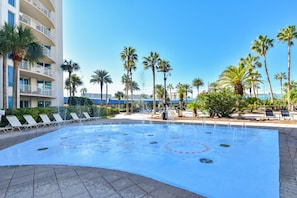 Une piscine dotée de jets d’eau, entourée de palmiers et de chaises longues, avec un bâtiment en arrière-plan.
