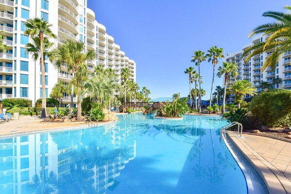 A hotel with a large swimming pool surrounded by palm trees and a multi-story building with balconies.