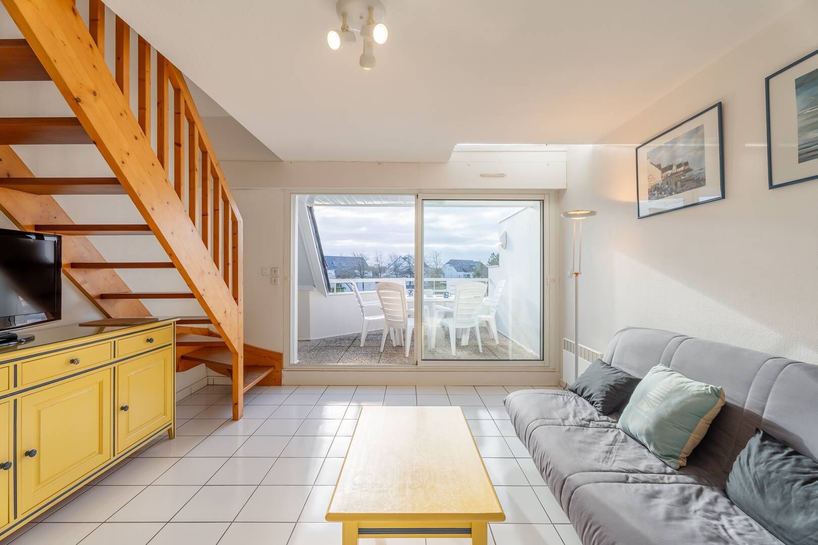 Een woonkamer met een houten trap, een bank, een geel kastje en een televisie.